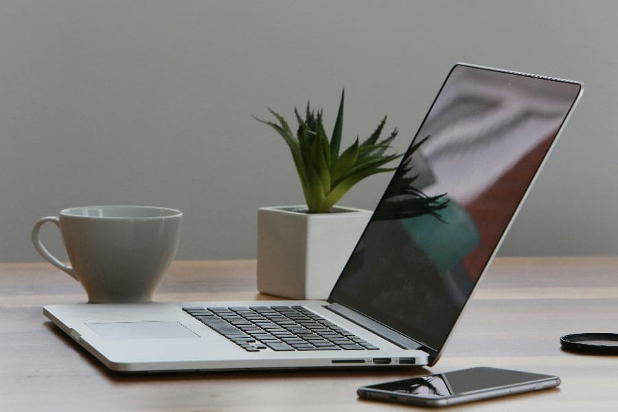 Arbeitsplatz mit IT-Leistungen, einschließlich Laptop und Smartphone auf einem hellen Tisch neben einer Kaffeetasse und einer Topfpflanze